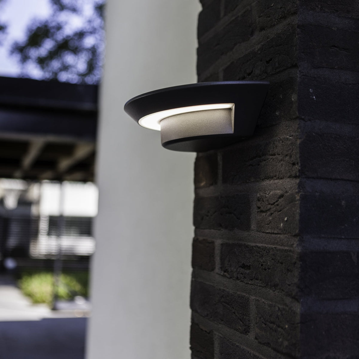 Modern black outdoor wall light with a curved design, emitting a soft glow, mounted on a dark brick pillar.