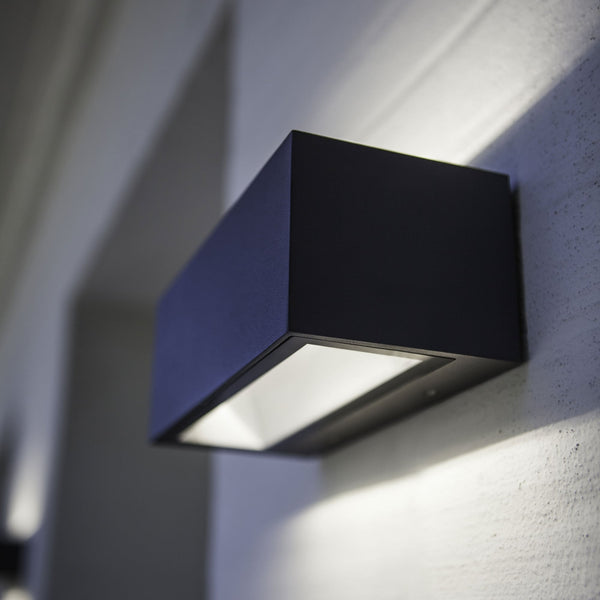 A dark grey outdoor wall light with a rectangular shape, illuminating a white wall.