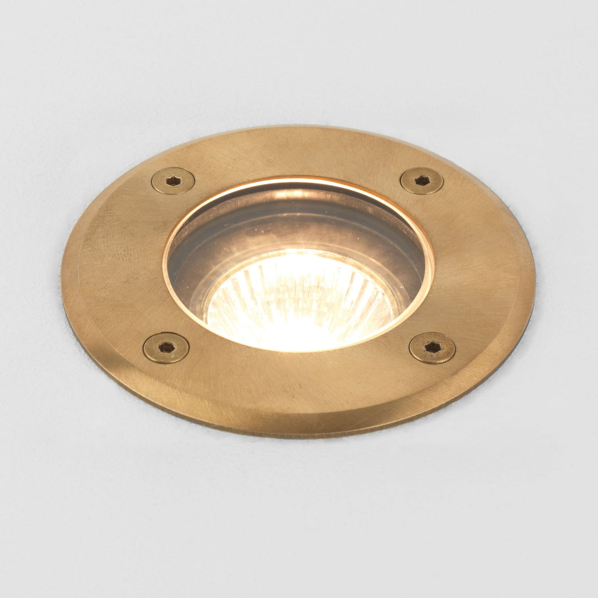 Round brass ground light with a clear glass cover and four visible screws, emitting a warm white light