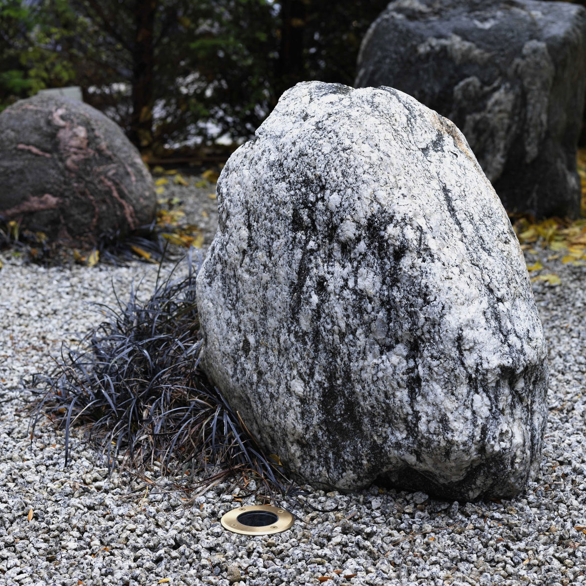 Large white granite boulder with dark streaks on a bed of small grey pebbles, with dark foliage behind
