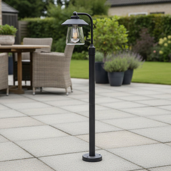 A black outdoor lamp post with a clear glass lantern and an Edison bulb, standing on a paved patio.