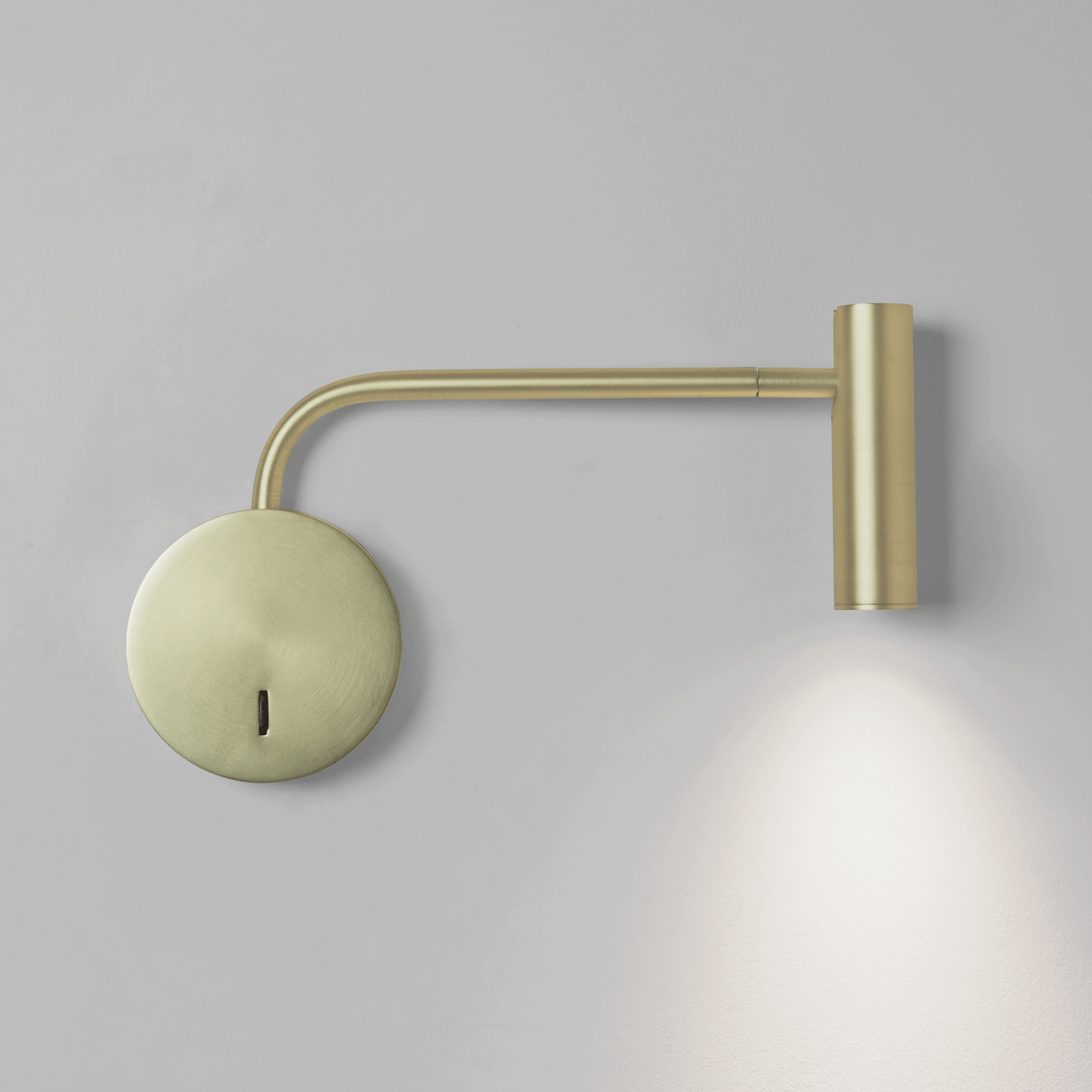 Brass wall lamp with a circular base and an adjustable arm, casting a downward light on a light grey wall