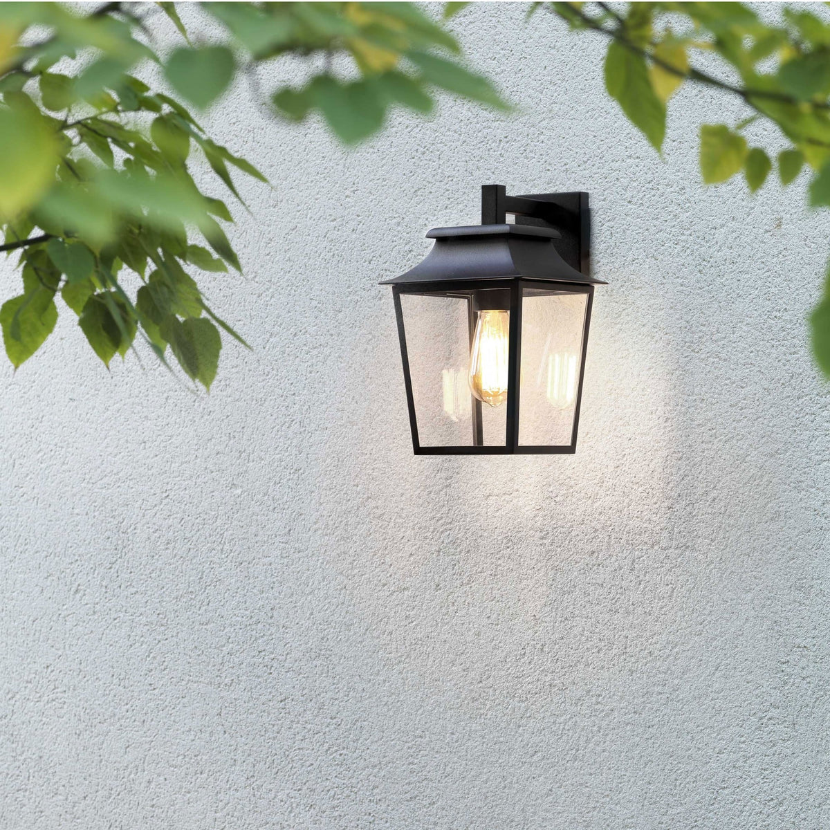 Black outdoor wall lantern with a clear glass shade and exposed filament bulb on a textured white wall