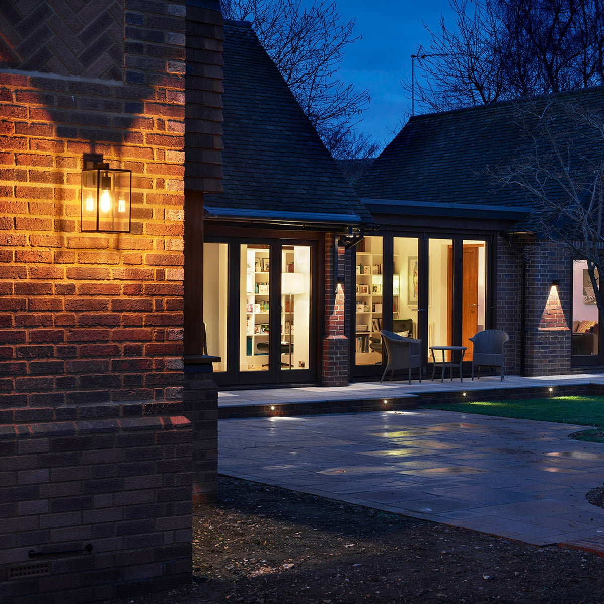 Exterior of a house at dusk with a wall light and ground lights illuminating a patio and garden