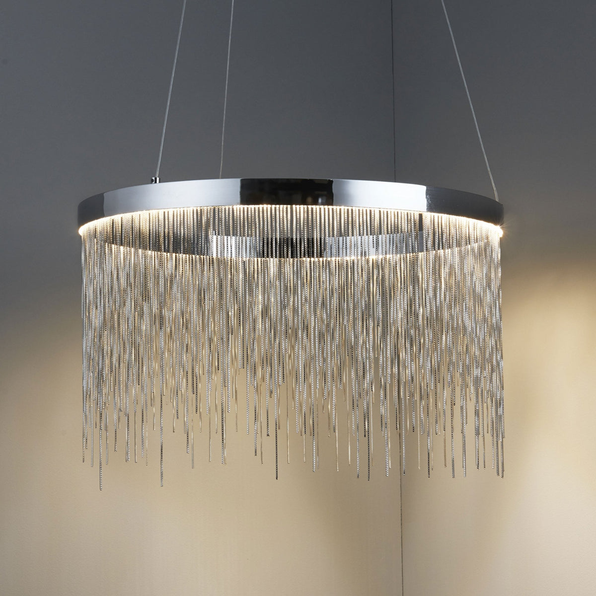 Chrome chandelier with cascading metal chains, illuminated from within, casting a warm glow in a dimly lit corner.