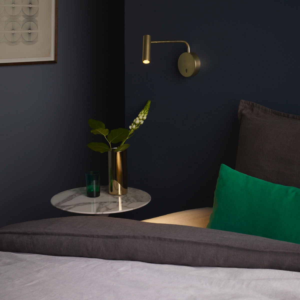A brass wall light illuminates a bedside table with a vase of flowers and a green glass