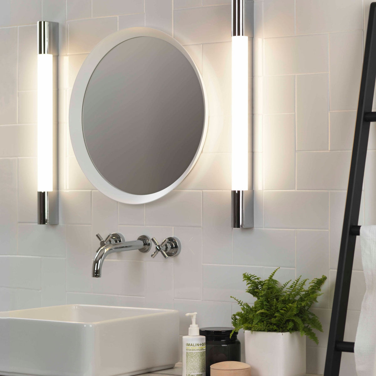 Bathroom with white subway tiles, a round mirror, two vertical chrome lights, a sink, and a plant