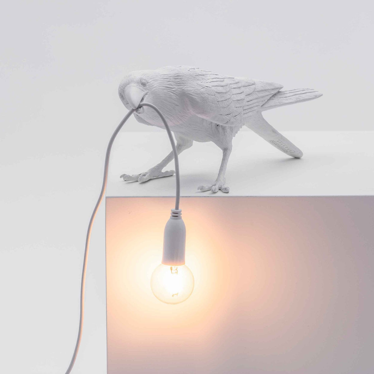 A white crow-shaped lamp with a lightbulb in its beak, positioned on a white ledge.