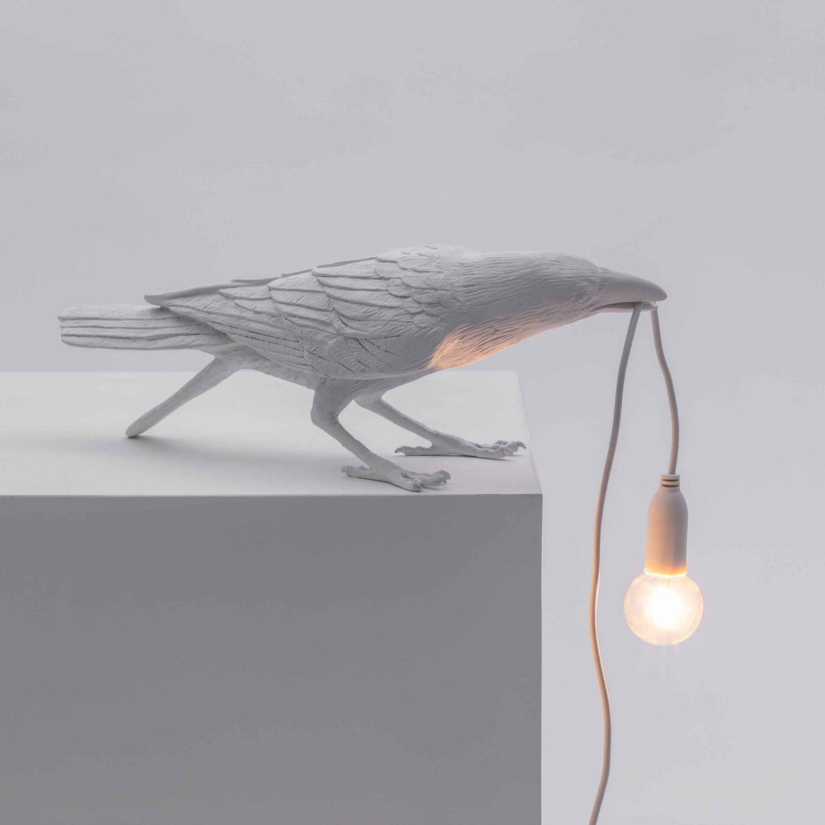 A white crow-shaped lamp with a lightbulb in its beak, angled to the right on a white surface.