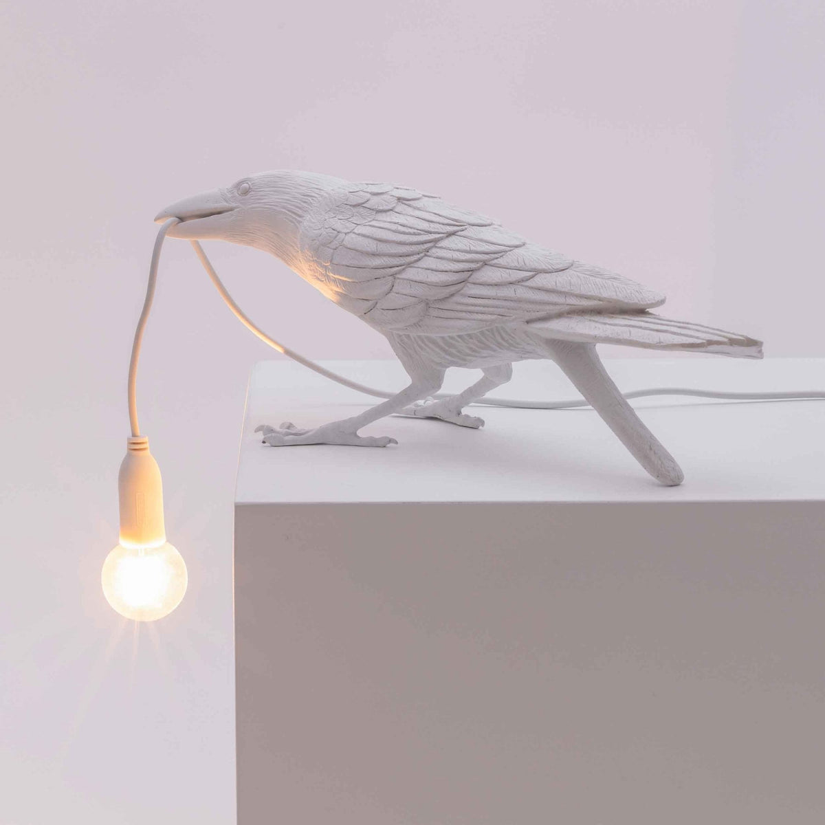 A white crow-shaped lamp on a white surface with a glowing lightbulb in its beak.