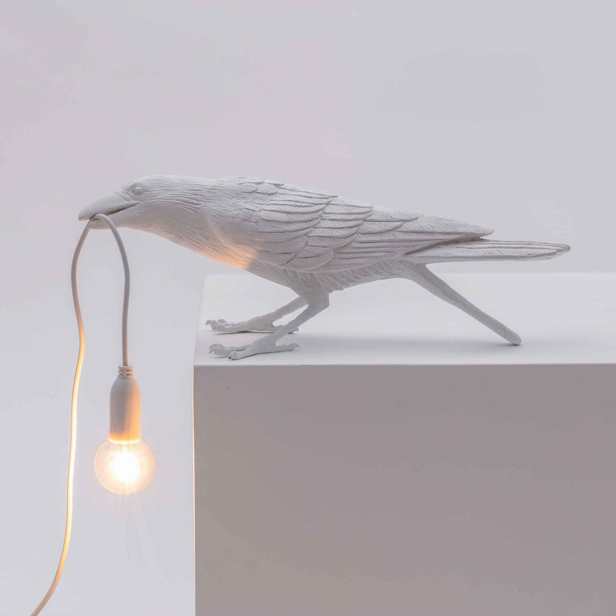 A white crow-shaped lamp on a white ledge, holding a glowing lightbulb in its beak.