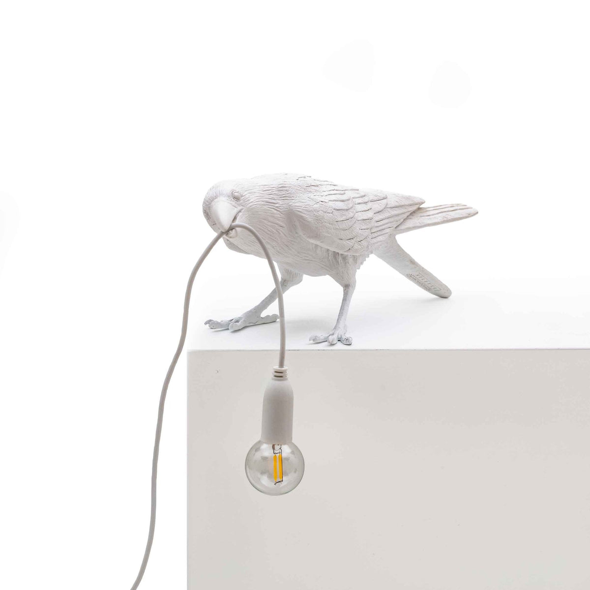 A white crow-shaped lamp with a lightbulb featuring a filament, on a white surface.