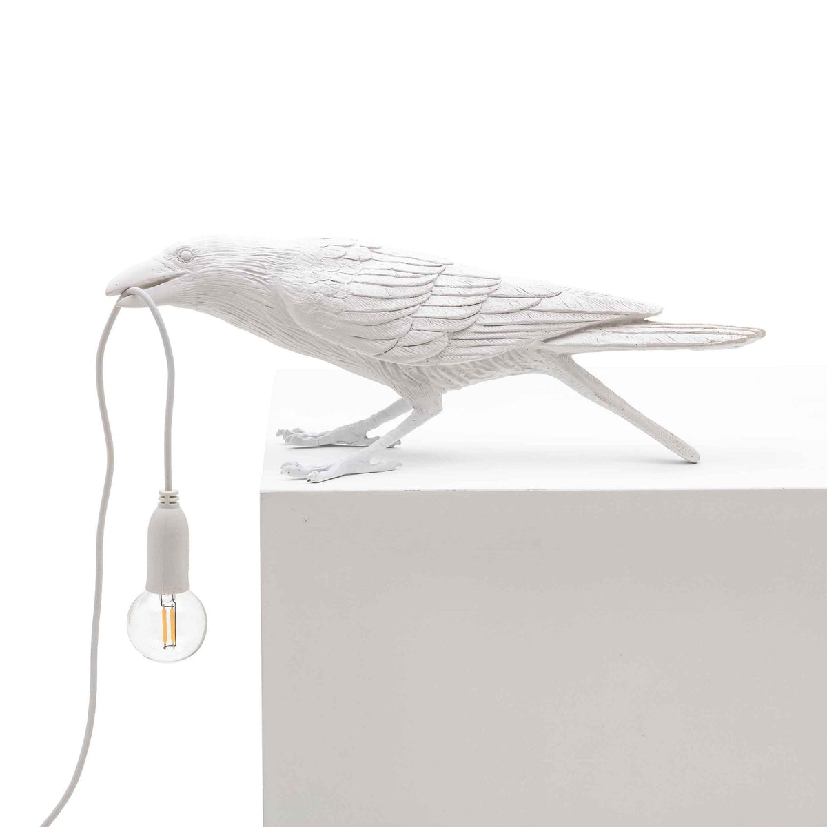 A white crow-shaped lamp with a lightbulb in its beak, on a white block, facing right.