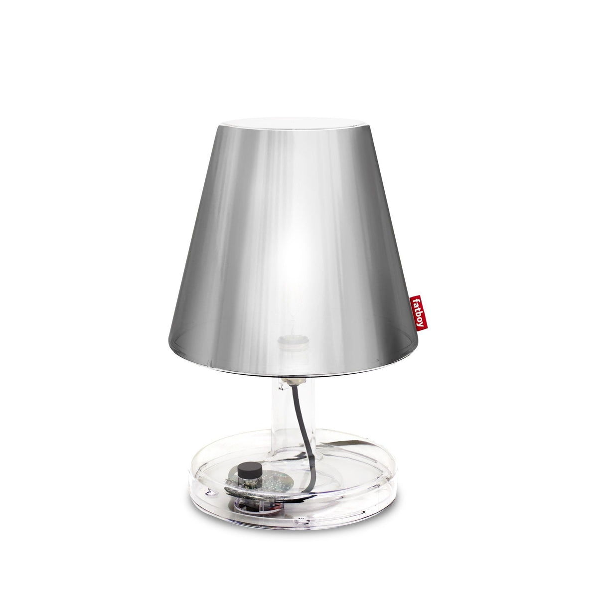 A brushed metal table lamp with a clear base and a red tag, illuminated against a white background.