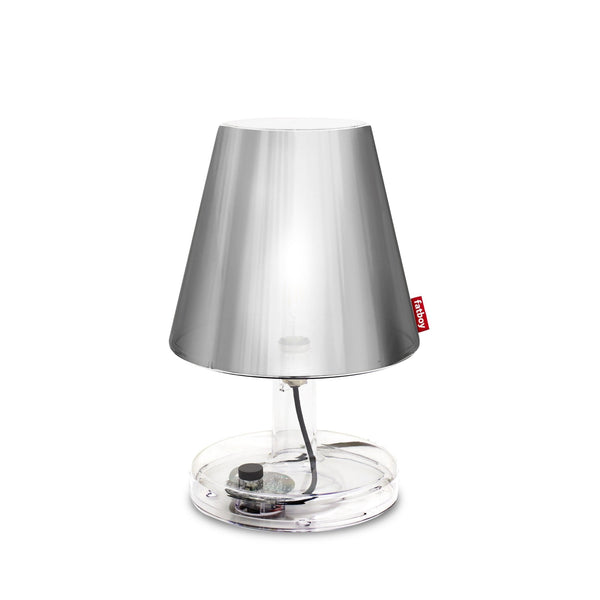 A brushed metal table lamp with a clear base and a red tag, illuminated against a white background.