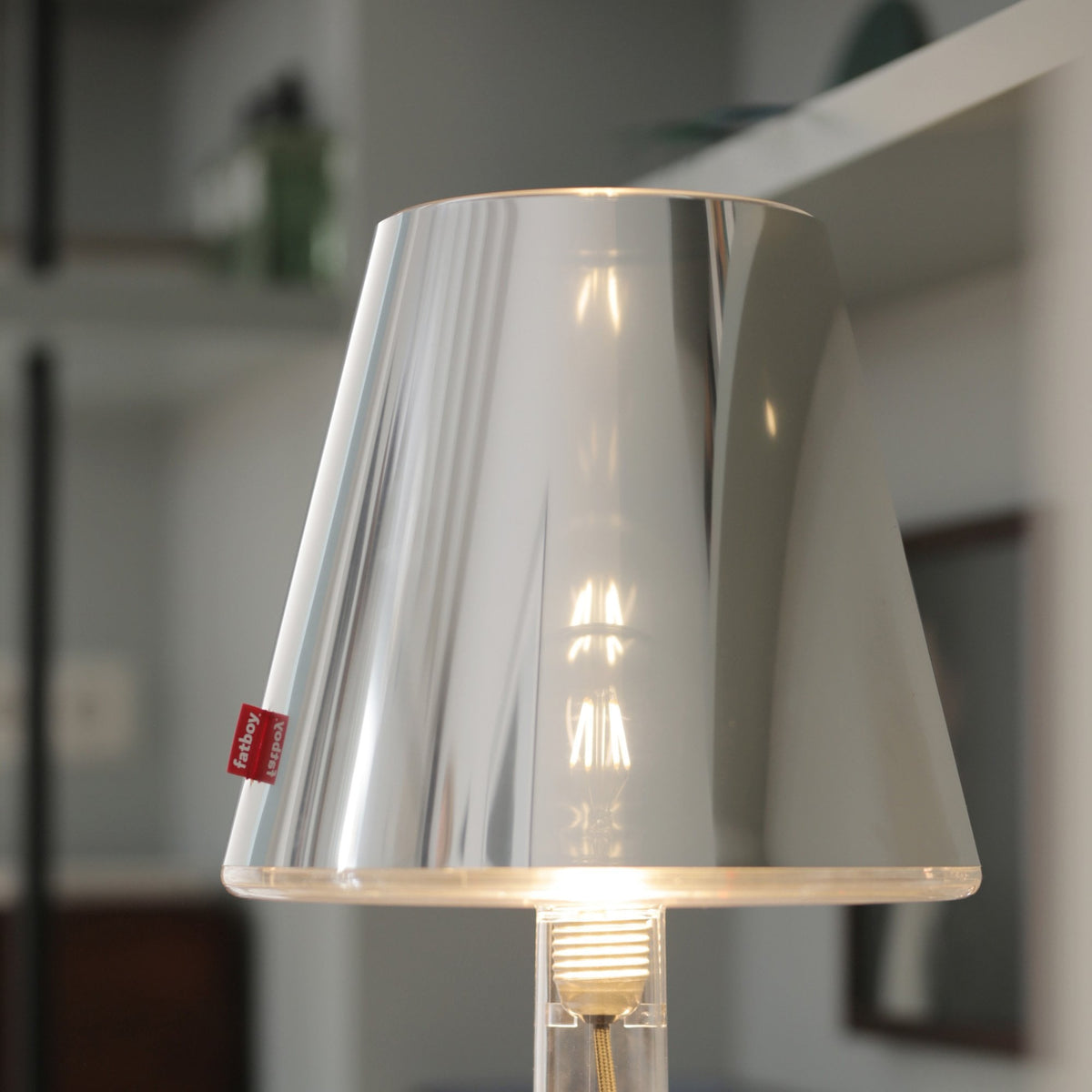 A close-up of the illuminated brushed metal table lamp with a clear base, on a shelf.