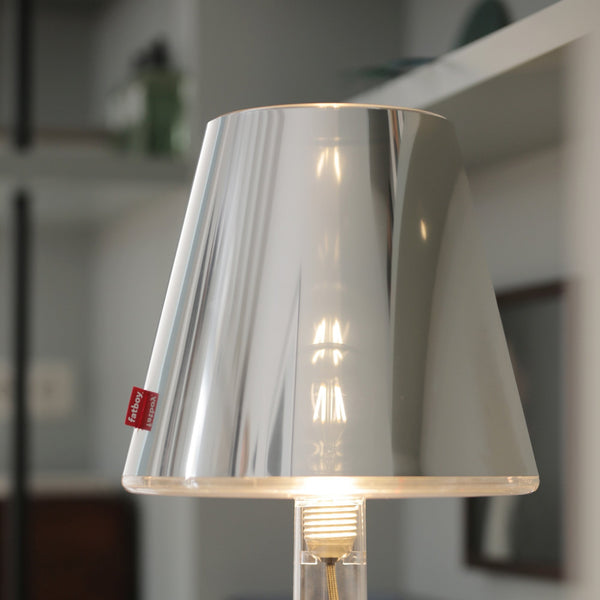 A close-up of the illuminated brushed metal table lamp with a clear base, on a shelf.