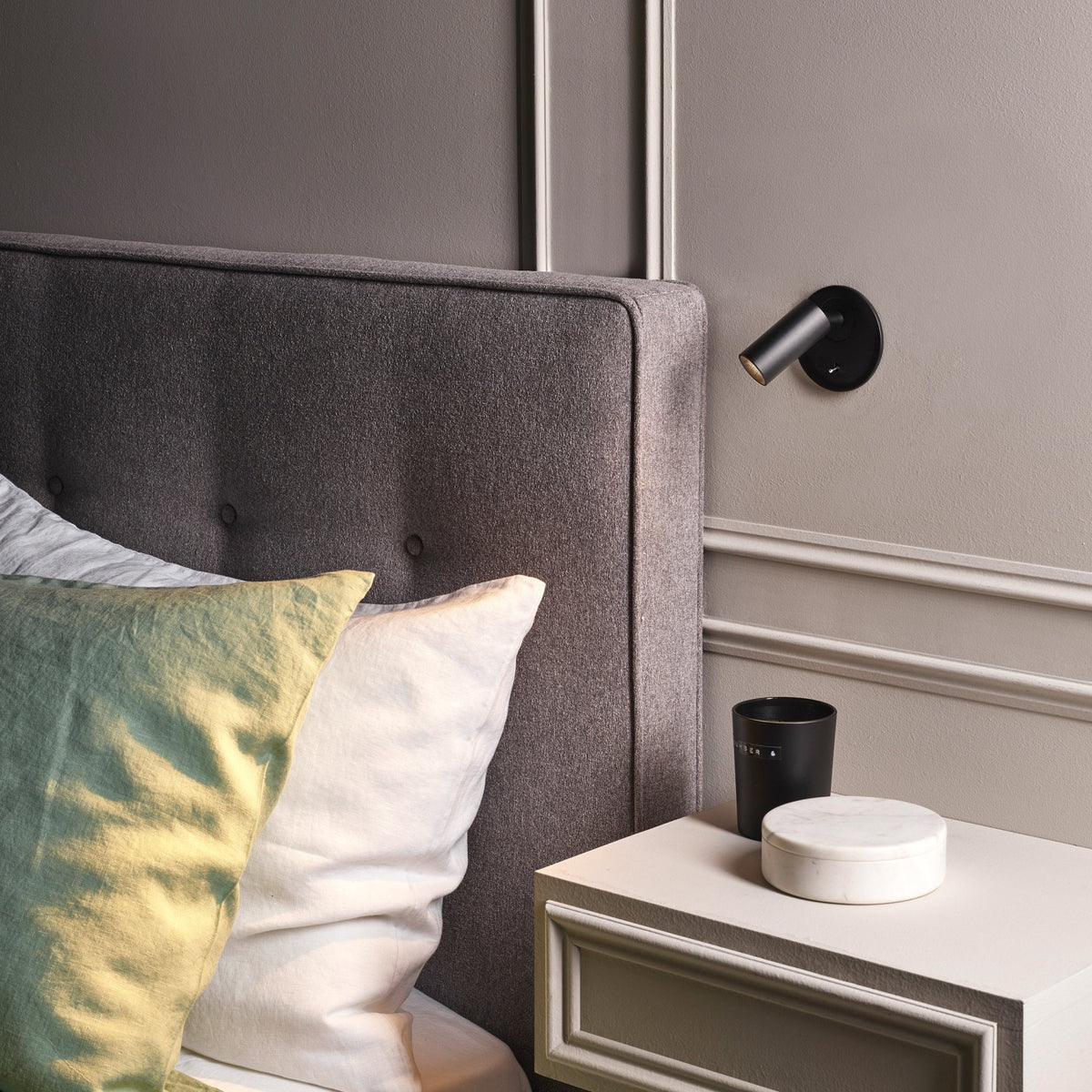 Bedroom with grey upholstered headboard, black wall light, and white bedside table with marble box