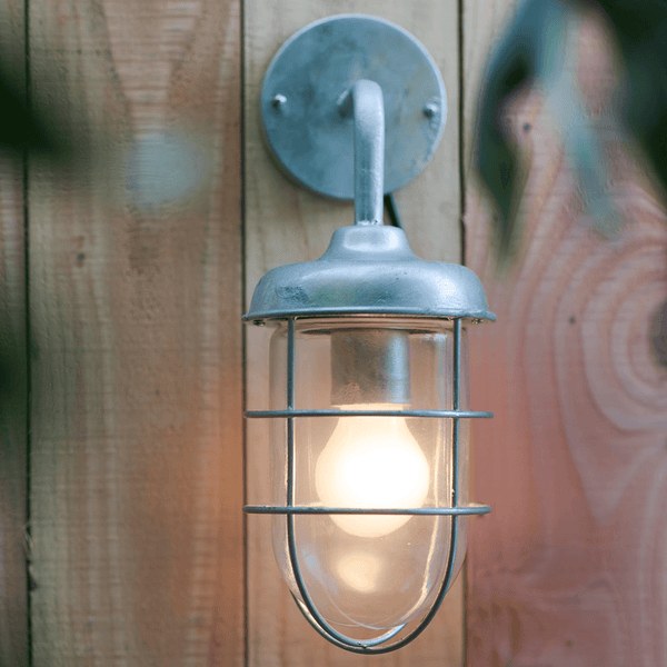 A close-up of a caged outdoor wall light with an exposed bulb, fixed to a wooden fence.