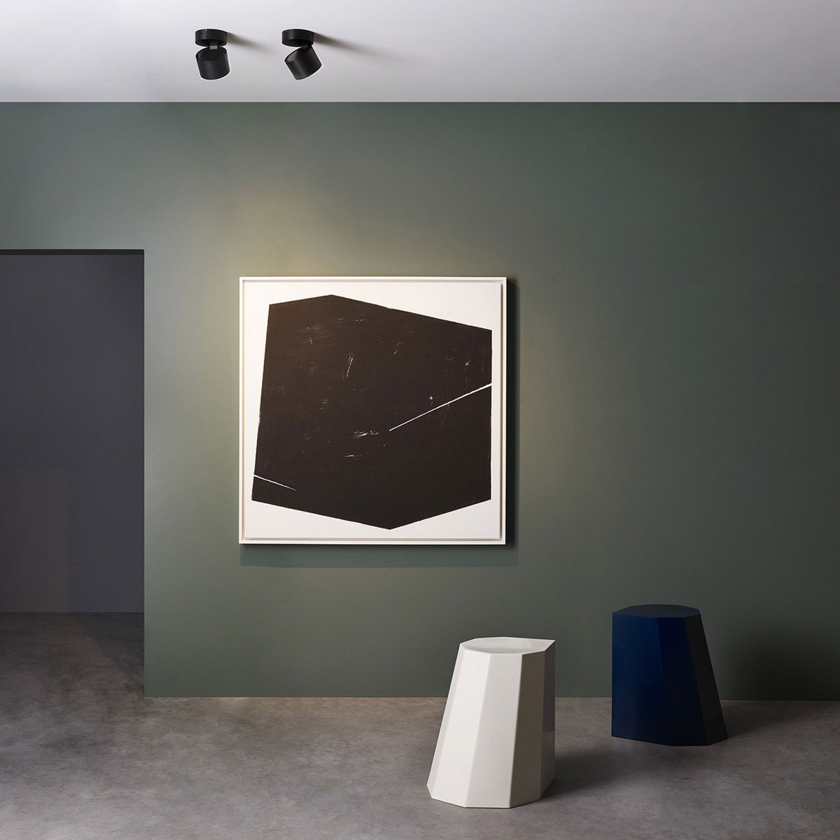 Two black ceiling spotlights illuminate a framed abstract black and white painting on a green wall