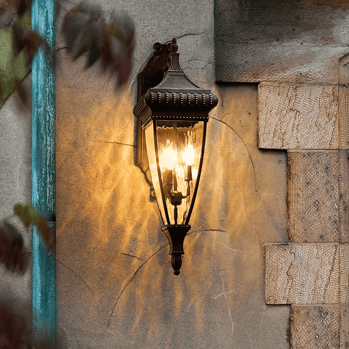 An ornate bronze outdoor wall light casting a glow on a textured wall.