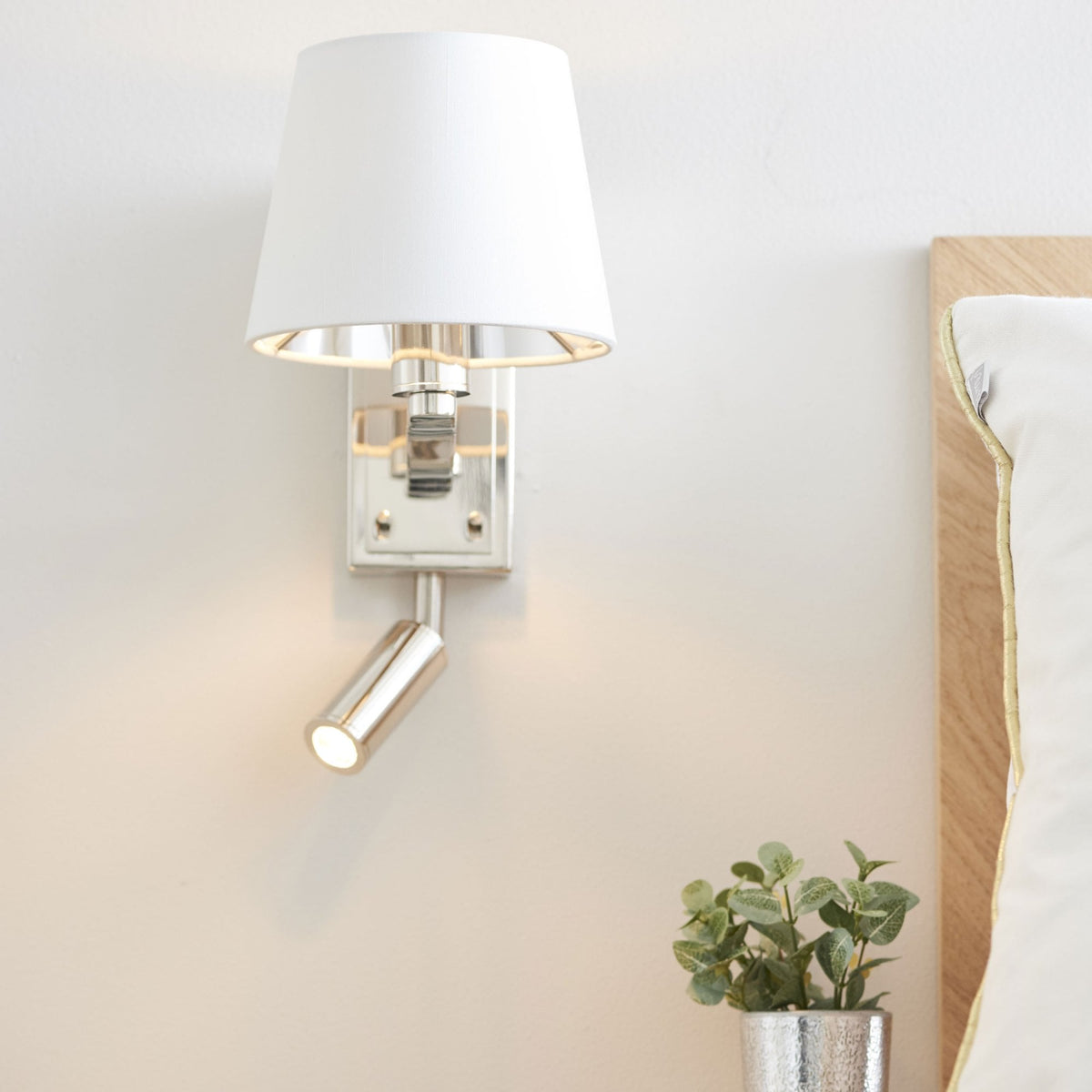Polished nickel wall light with a white fabric shade and an adjustable reading light, next to a bed.