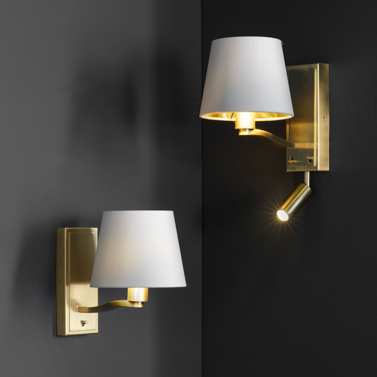 Two brass wall lights with white shades and gold interiors, one with an illuminated spotlight, on a dark corner wall.