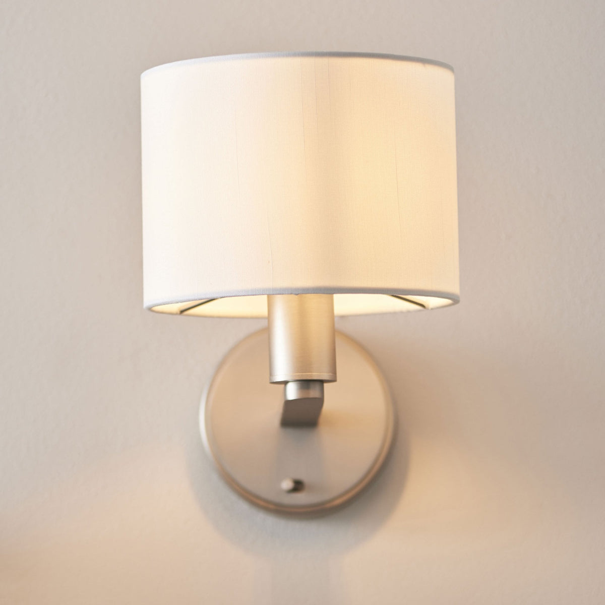 Wall-mounted lamp with a white cylindrical shade and brushed nickel base, casting a warm glow on a light wall.