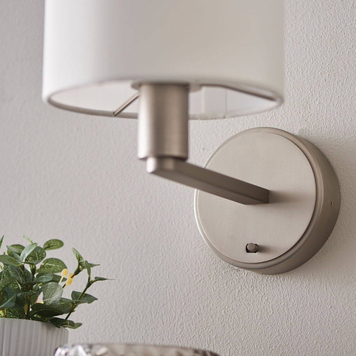 Wall-mounted lamp with a white cylindrical shade and brushed nickel base, with a green plant in the foreground.