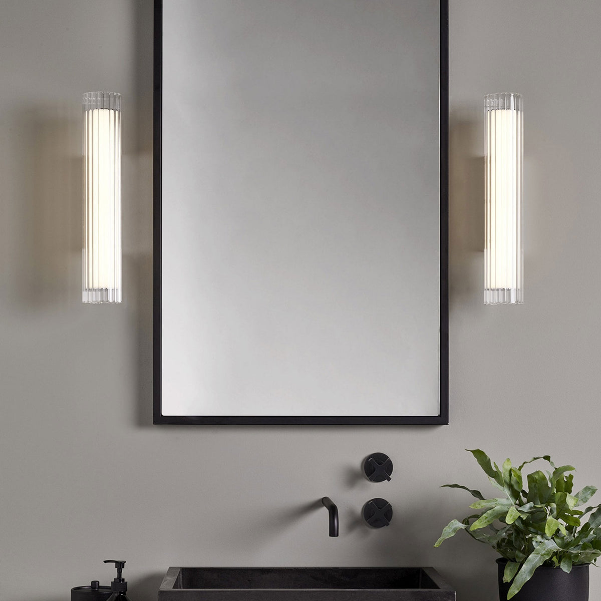 Two vertical fluted glass wall sconces flank a mirror above a black sink with a plant to the right