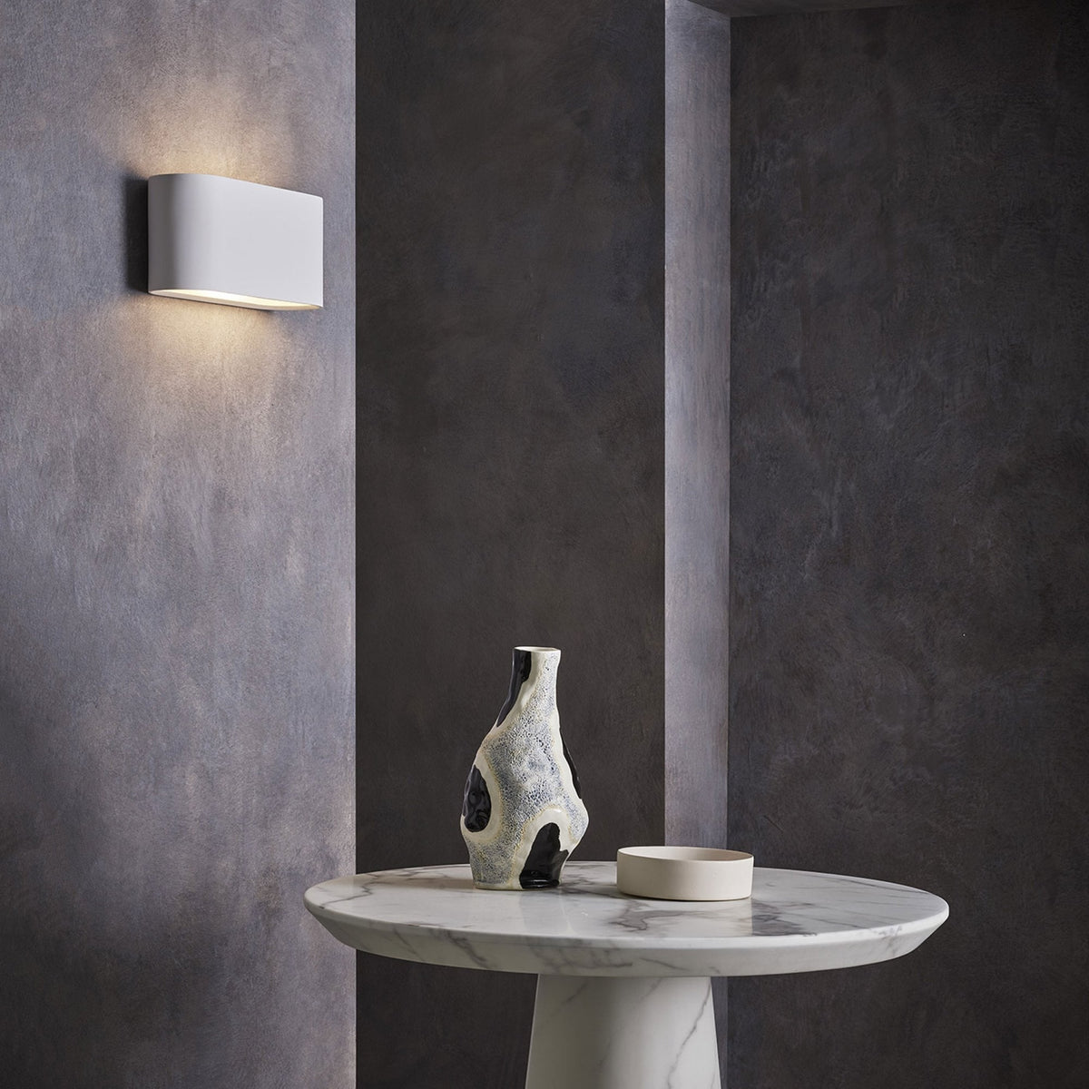White rectangular wall light illuminating a textured gray wall above a marble table with a decorative vase