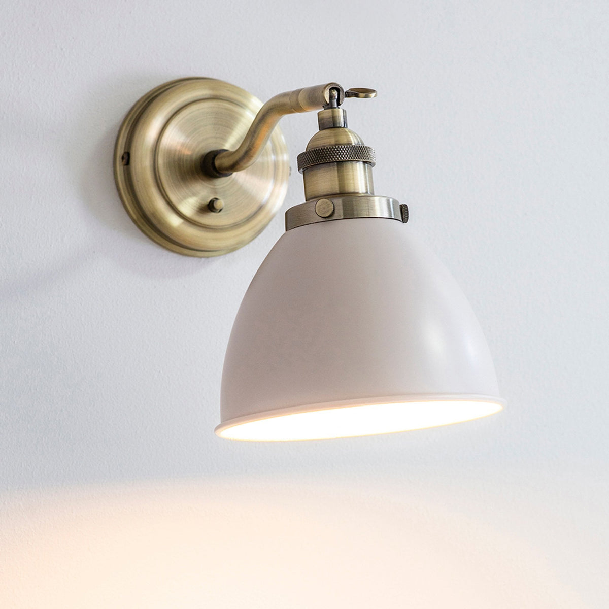 An antique brass wall light with a light grey dome shade, illuminating a white wall.