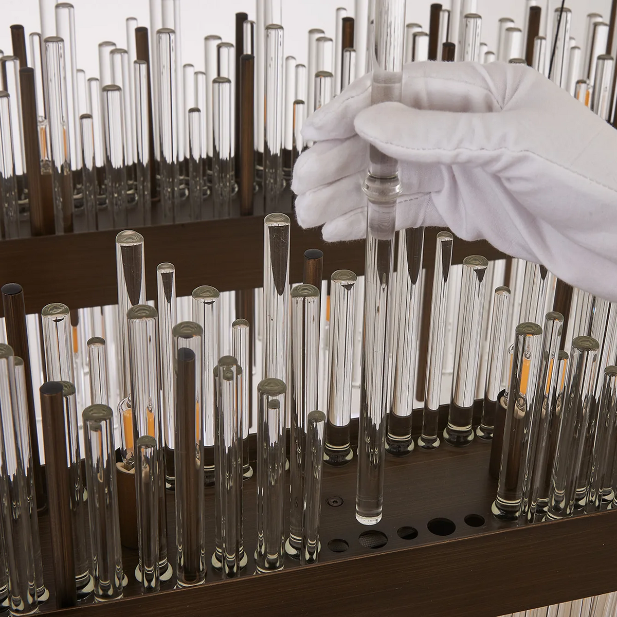Gloved hand placing a glass rod into a dark wooden base with many other glass and dark rods