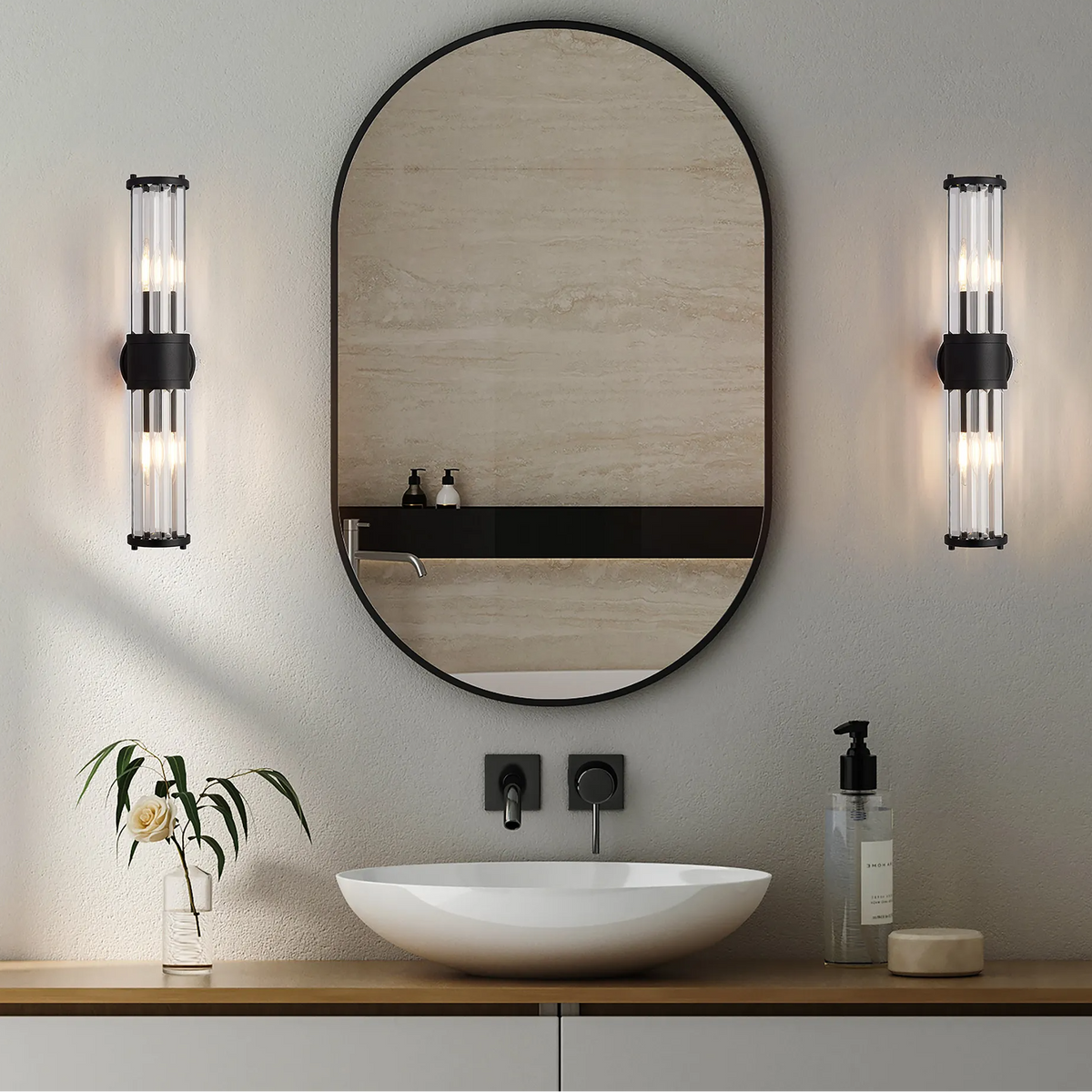 Two black and clear glass wall sconces flanking an oval mirror above a white sink and wooden vanity