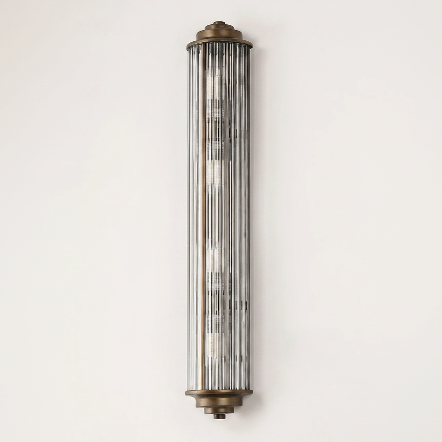 A vertical wall light with a bronze base and top, featuring a cylindrical glass shade with vertical fluting