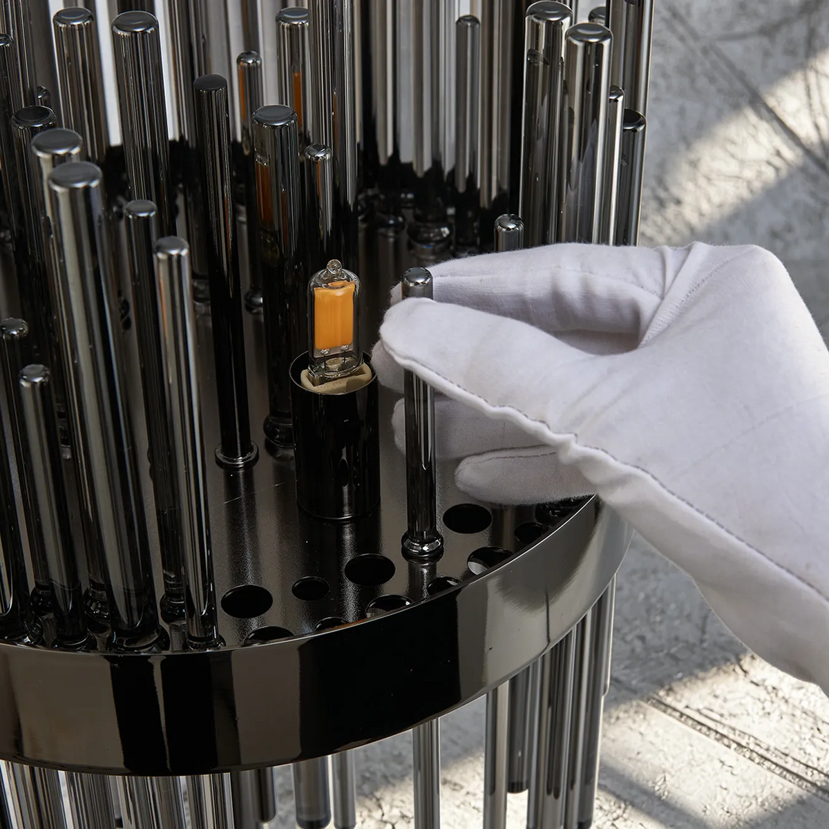 Gloved hand adjusting a metal rod on a black circular base with an orange light bulb