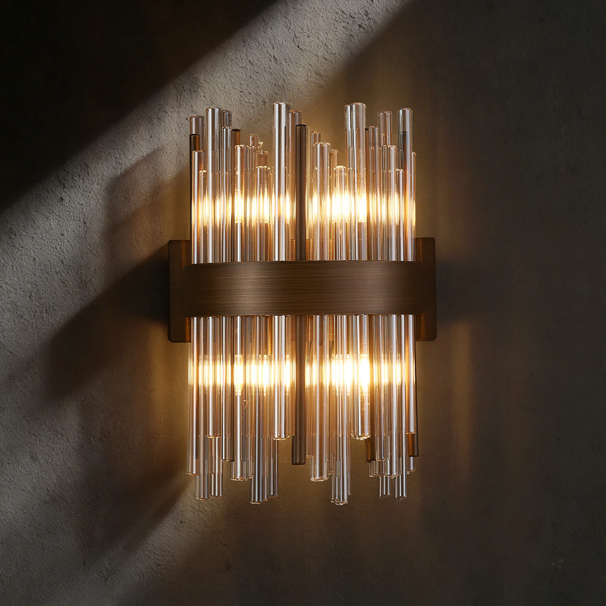Wall sconce with multiple vertical glass tubes and a horizontal bronze band, illuminated against a dark wall