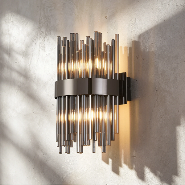 Wall sconce with multiple vertical glass tubes in varying lengths, held by a dark metal band, illuminated