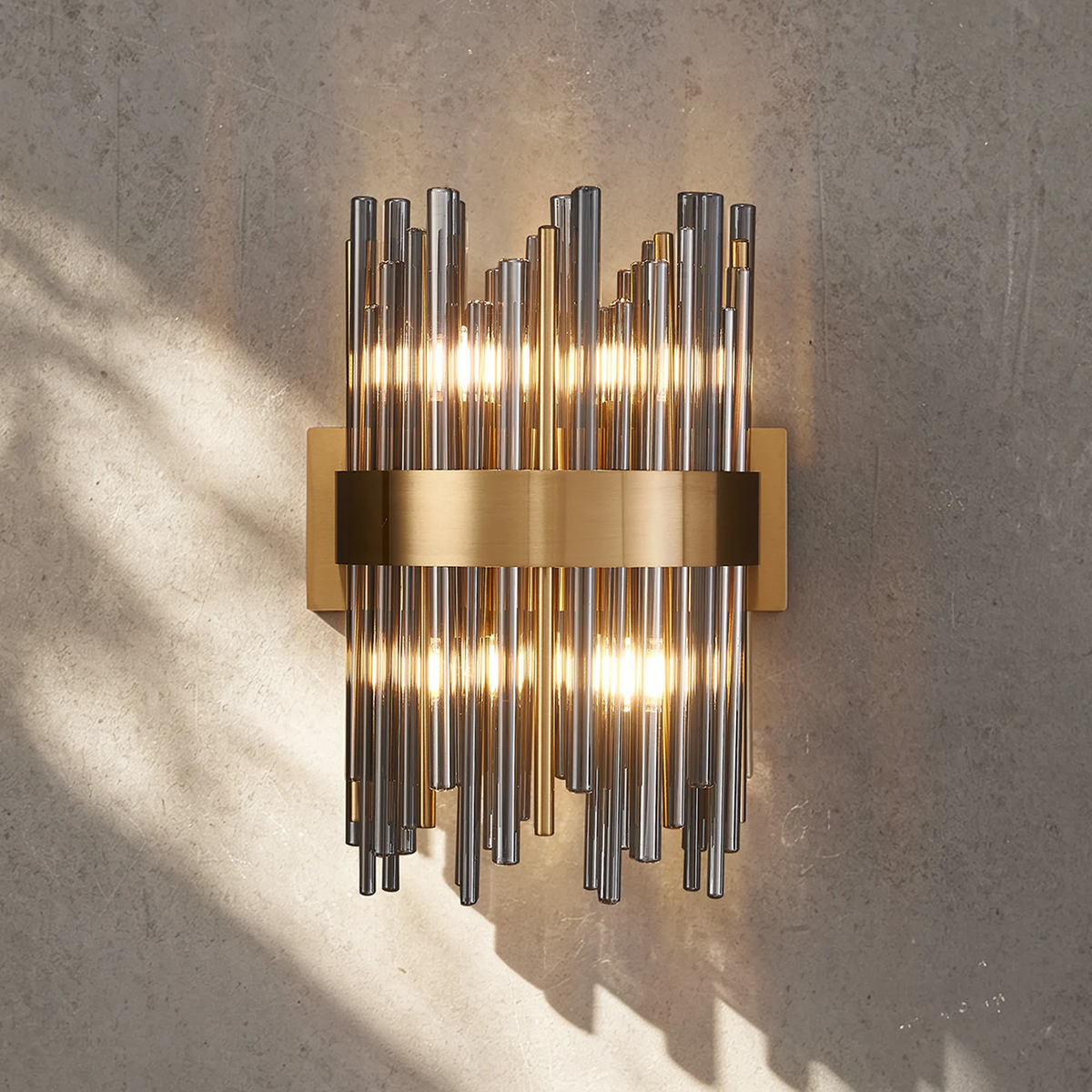 Wall sconce with vertical glass rods and a horizontal brass band, illuminated against a textured wall