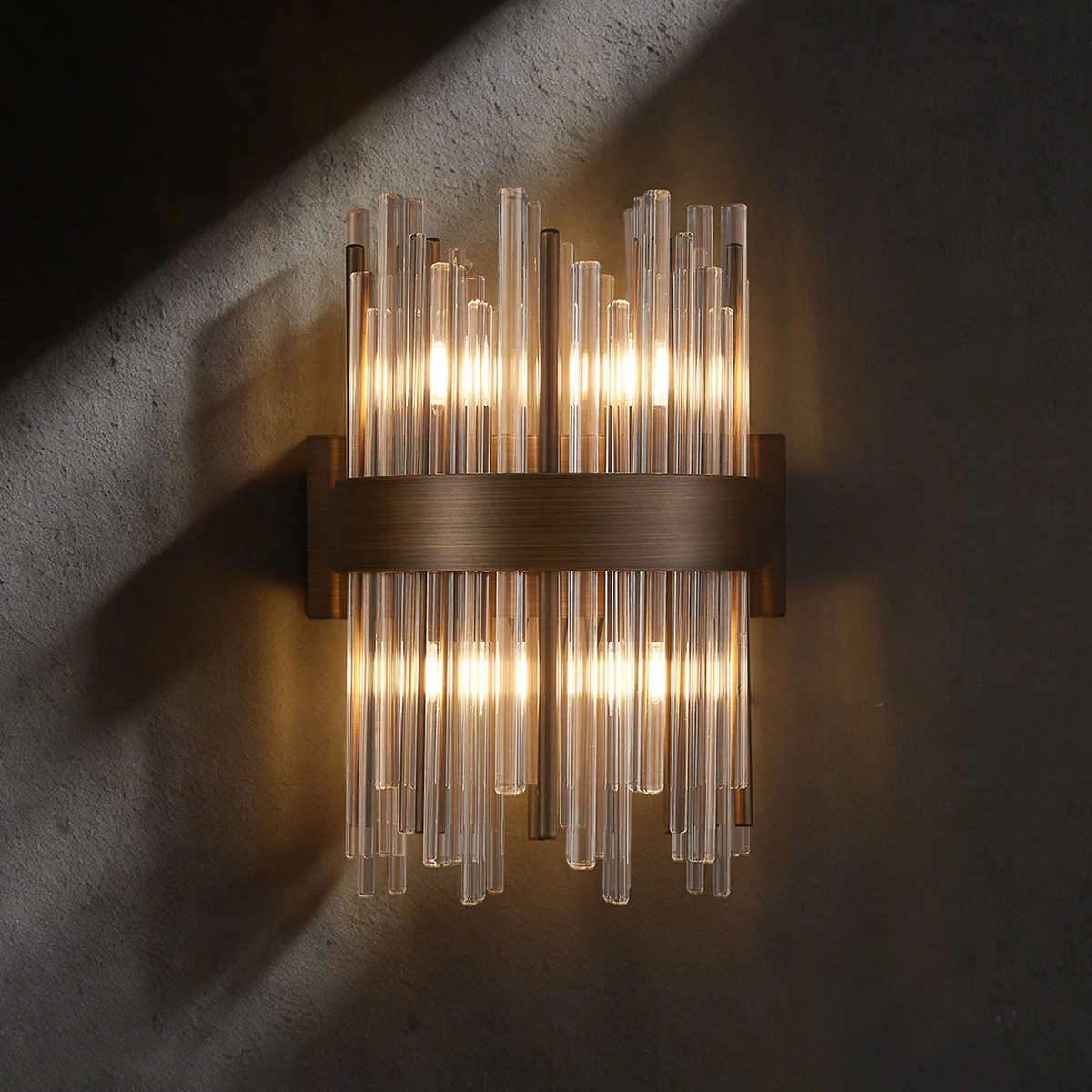 Modern wall sconce with vertical glass rods and a bronze band, illuminated against a dark textured wall