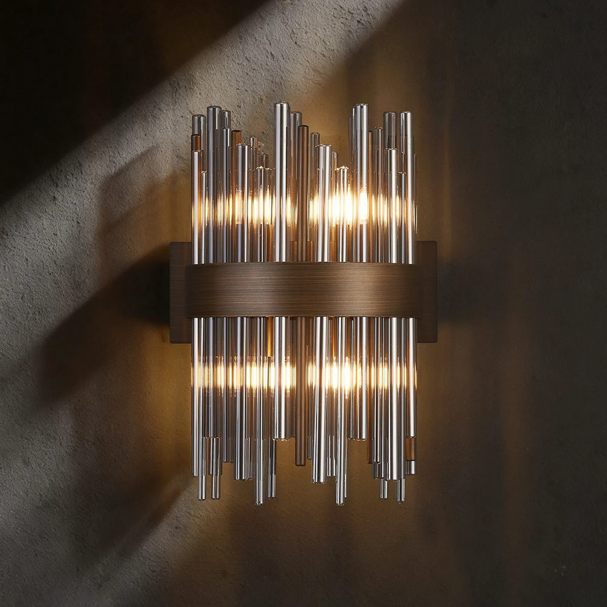 Wall sconce with vertical glass rods and a horizontal bronze band, illuminated against a dark wall