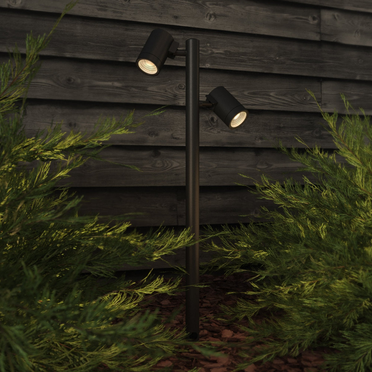 Two black outdoor spotlights on a pole, illuminating green bushes in front of a dark wooden fence