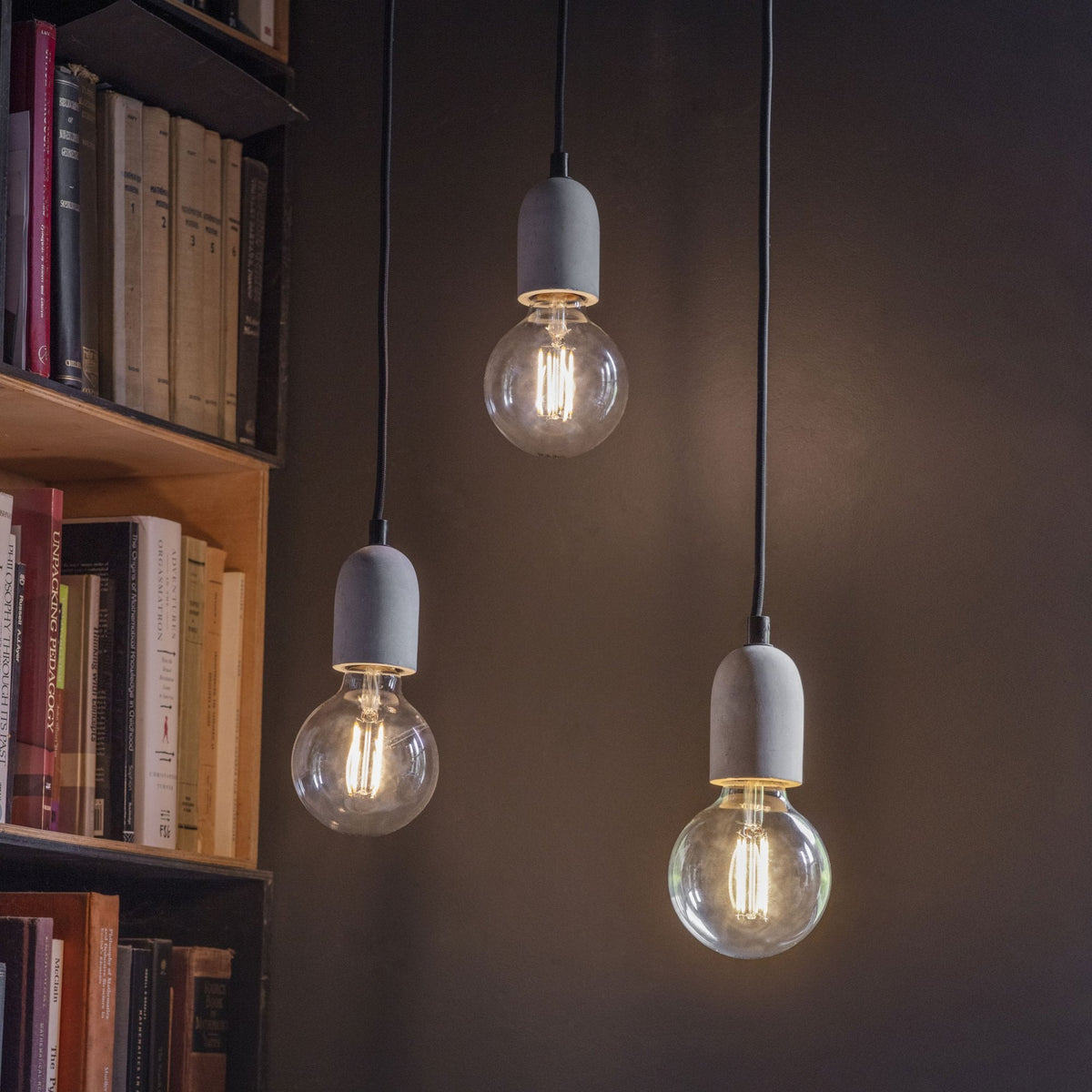 Three hanging light bulbs with concrete bases illuminate a dark room next to a bookshelf filled with books.