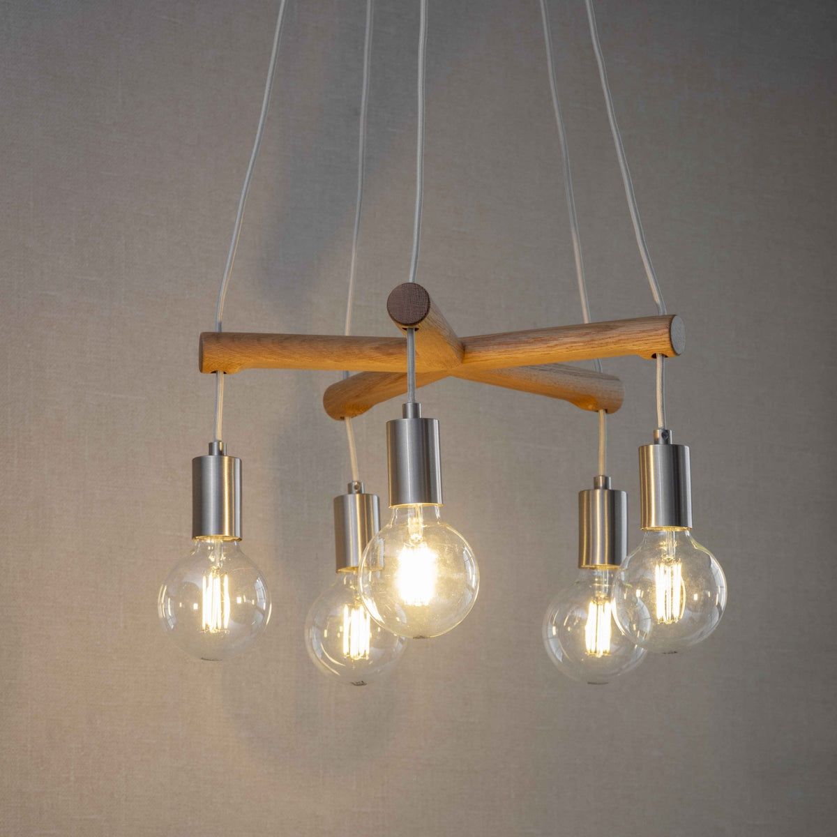 A modern chandelier with five exposed bulbs hanging from a wooden crossbar, against a neutral background.