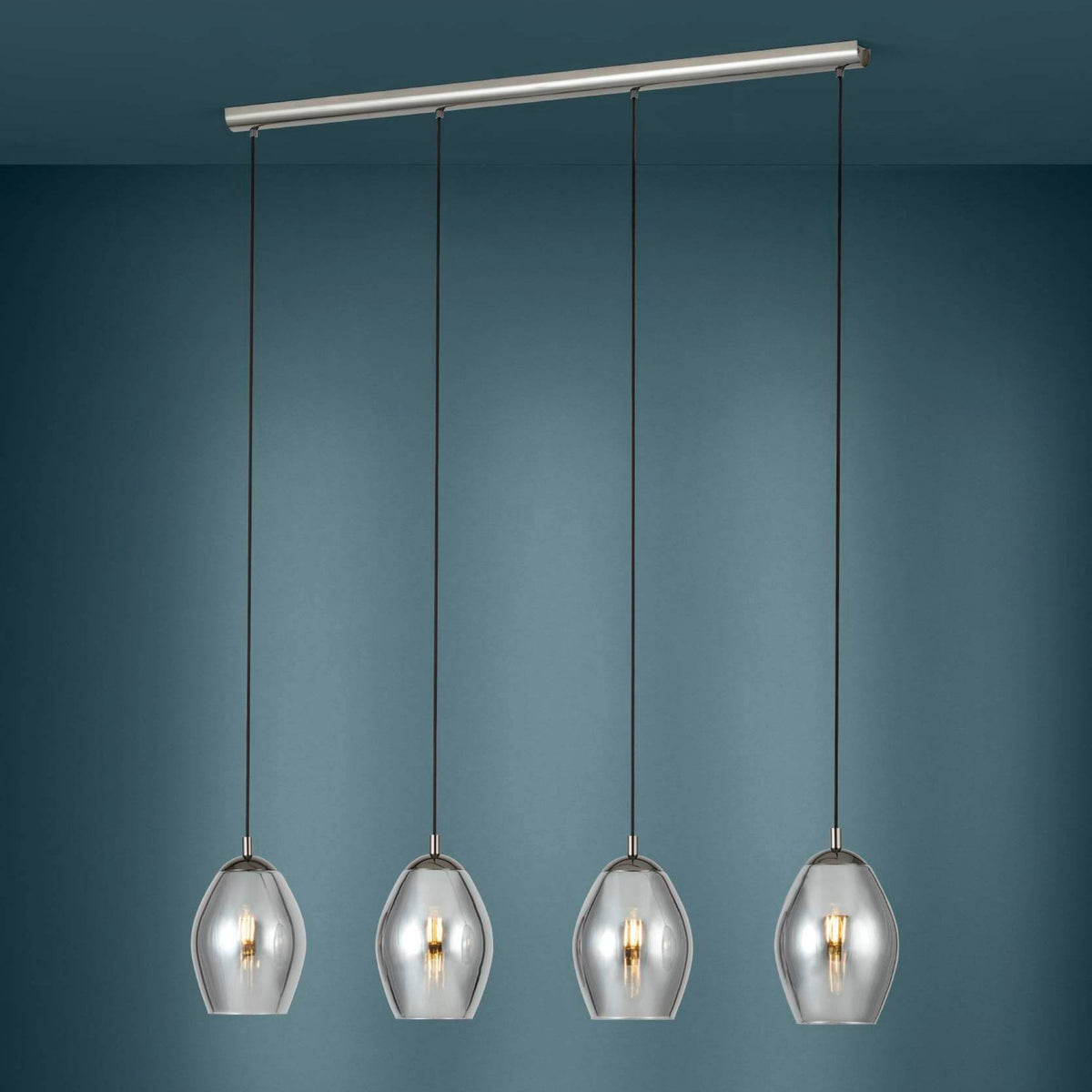 Four clear glass pendant lights with warm bulbs hang from a brushed metal bar against a dark blue wall.