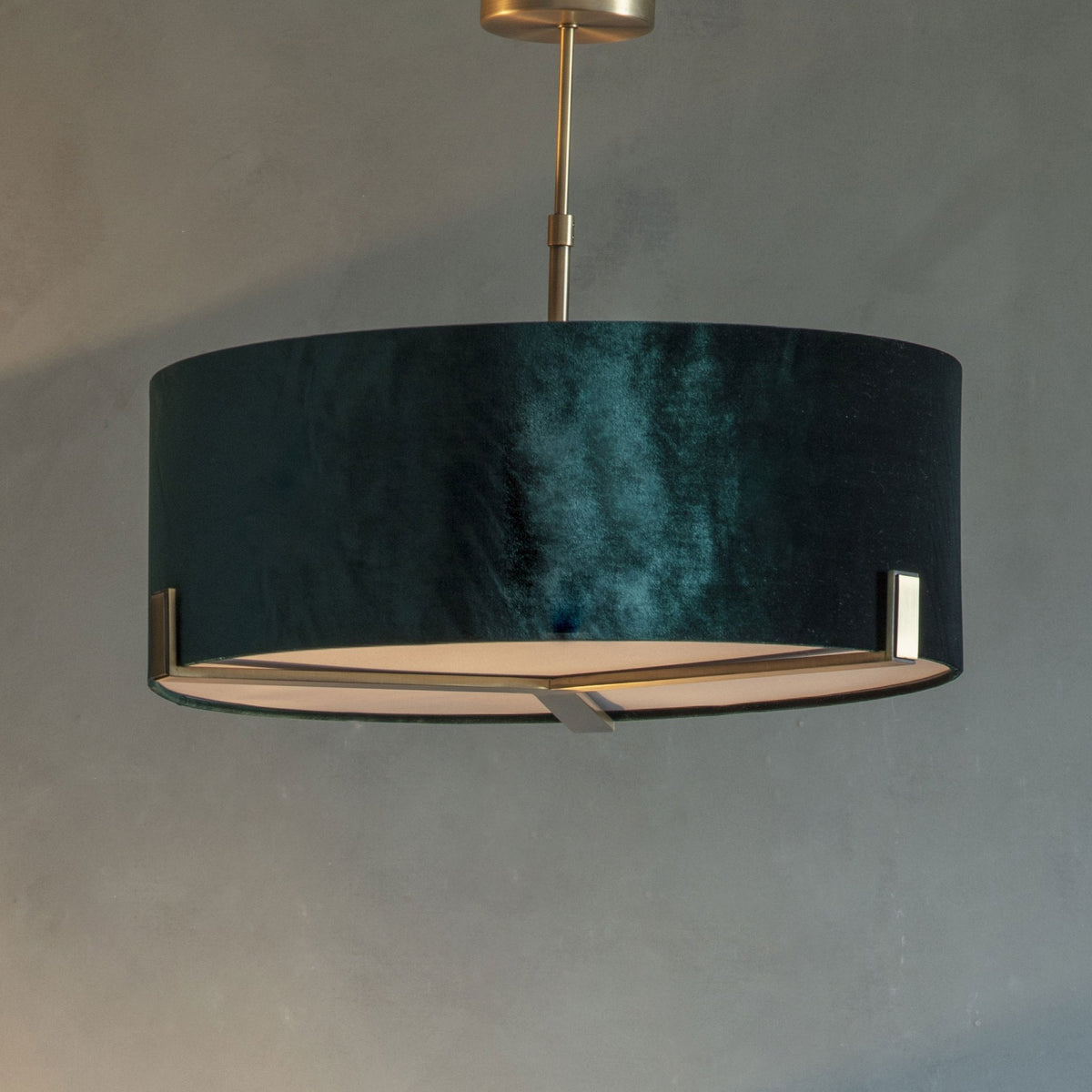 A close-up of a dark green velvet drum pendant light with gold accents against a gray wall.