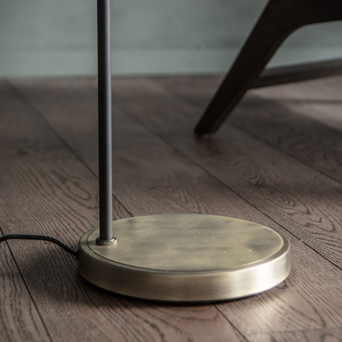 Close-up of the round brass base of a floor lamp on a wooden floor next to a chair leg.