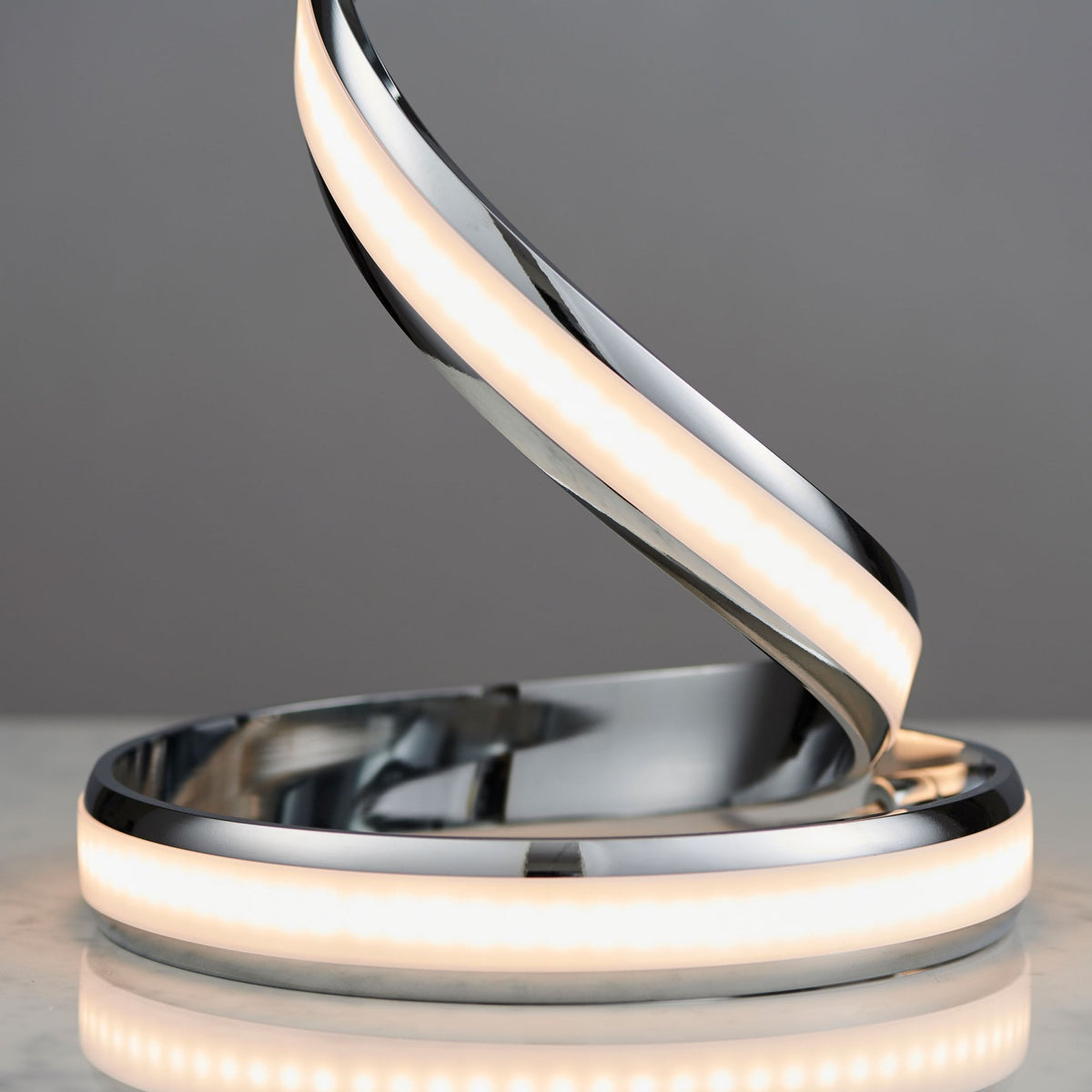 Close-up of the base of a chrome spiral LED table lamp, showing the illuminated warm light strip and its reflection.