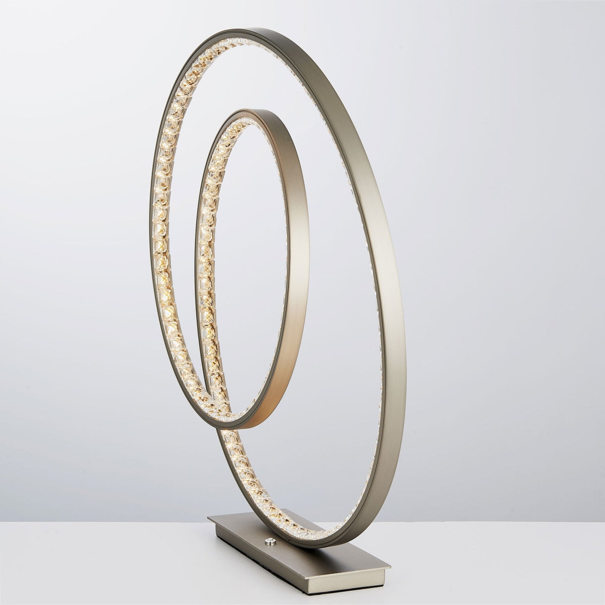 Modern table lamp with two intertwined rings, one illuminated with crystals, on a rectangular base.