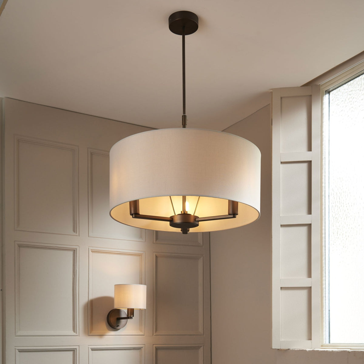 Drum pendant light with a white shade and dark bronze accents hanging in a room with paneled walls.